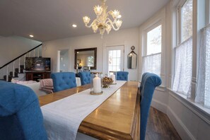 A dining area with a long wooden table, blue upholstered chairs, a chandelier, and large windows.