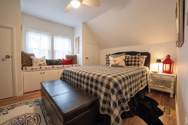 A bedroom with a bed, a bench, a nightstand, a window with curtains, and a ceiling fan.