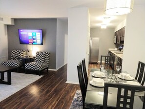 A modern living room with a flat-screen TV, a dining area with a glass table, and a kitchen in the background.