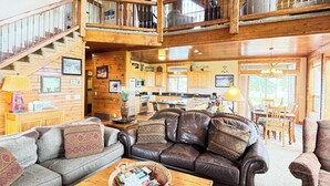 A spacious living area with a high ceiling, wooden staircase, and a kitchen in the background. - 35 MI to YNP | Private Hot Tub | Waterfront Access (Island Park)