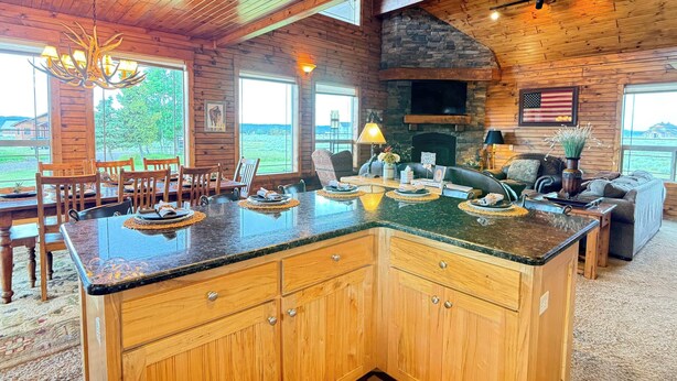 A wooden cabin interior with a large kitchen island, dining area, and a stone fireplace. - 35 MI to YNP | Private Hot Tub | Waterfront Access (Island Park)