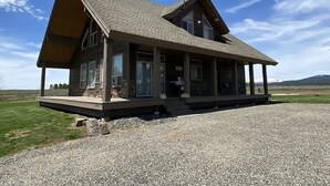 A two-story log cabin with a covered porch, large windows, and a gravel driveway. - 35 MI to YNP | Private Hot Tub | Waterfront Access (Island Park)