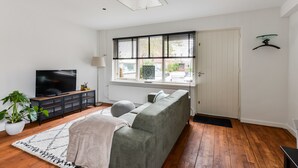 Ein modernes Wohnzimmer mit grauem Sofa, einem Fernseher, einer Topfpflanze und einem großen Fenster mit Jalousien.