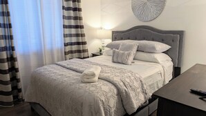 A neatly made bed with a grey headboard, a bedside table with a lamp, and a wall-mounted decorative piece. - Reflections - 2bdr condo in Central Raleigh (Raleigh)