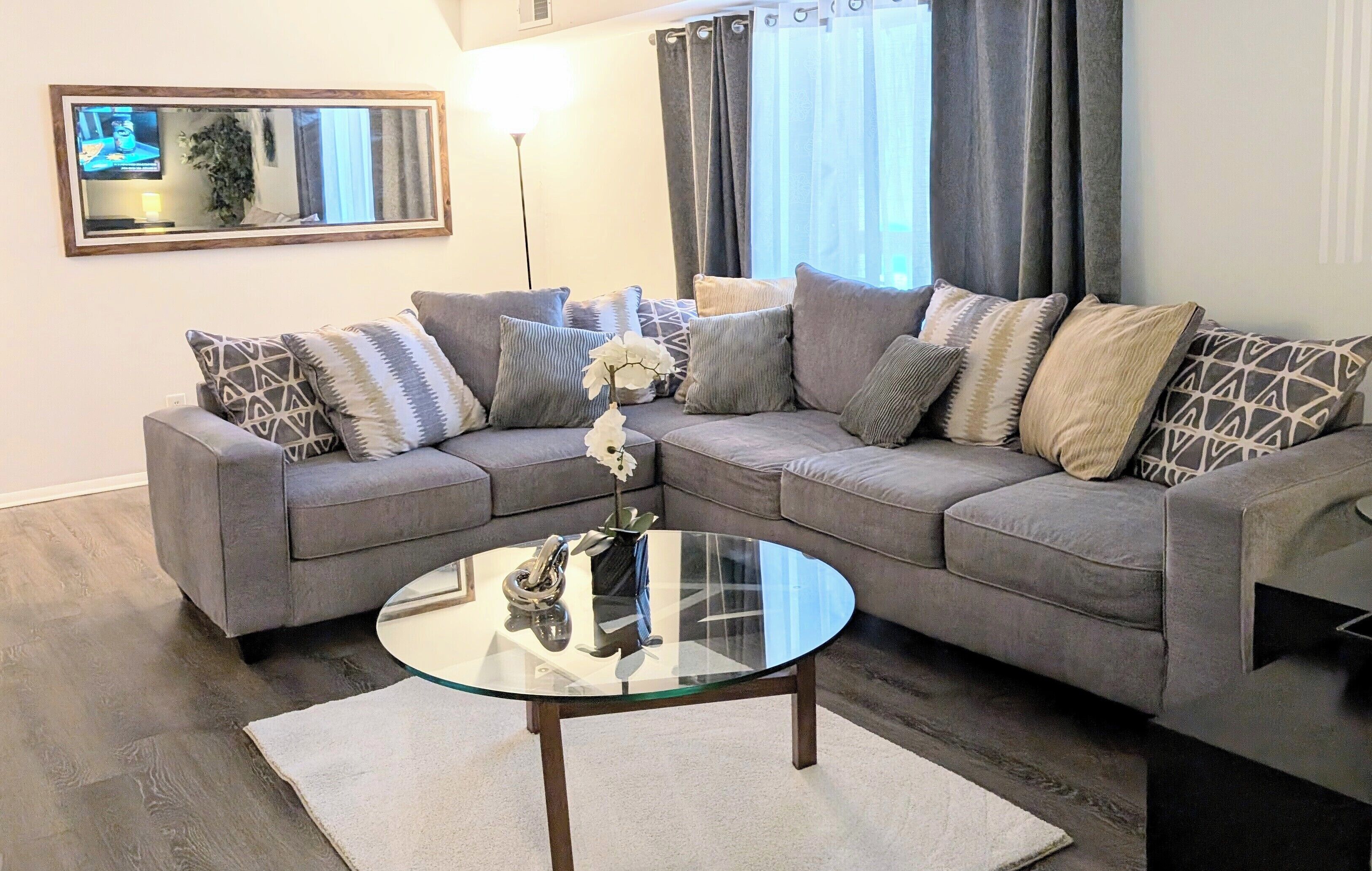 A modern living room with a grey sectional sofa, a glass coffee table, and a large mirror on the wall.