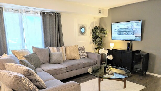 A living room with a grey sofa, a glass coffee table, and a TV on the wall. - Reflections - 2bdr condo in Central Raleigh (Raleigh)