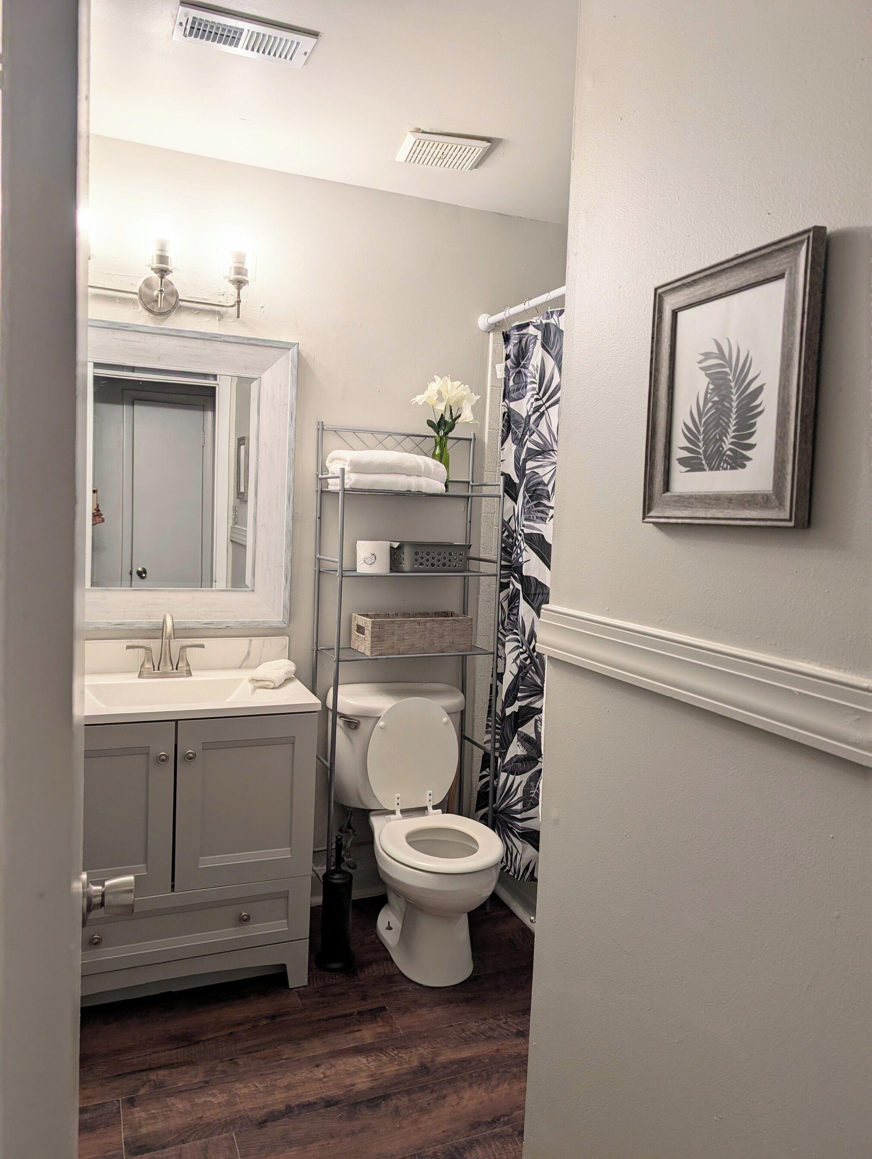 Une salle de bain comprenant des toilettes, un lavabo avec un miroir, un rideau de douche orné d’un motif de feuilles et un tableau encadré accroché au mur.