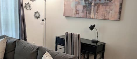 Un soggiorno con un divano grigio, una piccola scrivania con una lampada e un grande quadro astratto appeso al muro.