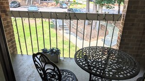 Ein Balkon mit einem Metalltisch und Stühlen, einem Topfpflanzen und Blick auf eine Straße mit parkenden Autos und Bäumen.