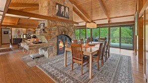 A spacious living area with a stone fireplace, wooden ceiling beams, a dining table with chairs, and large windows offering a view of greenery. - Frank Lloyd Wright-styled Mid-Century Modern Retreat on 5 Private Acres (Rabun Gap)