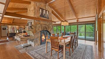 A spacious living area with a stone fireplace, wooden ceiling beams, a dining table with chairs, and large windows offering a view of greenery.