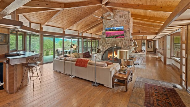 A spacious living area with a stone fireplace, wooden ceiling beams, and large windows offering a view of the outdoors. - Frank Lloyd Wright-styled Mid-Century Modern Retreat on 5 Private Acres (Rabun Gap)