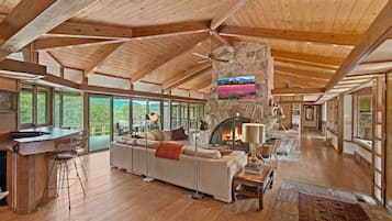 A spacious living area with a stone fireplace, wooden ceiling beams, and large windows offering a view of the outdoors.