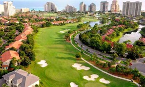 Un parcours de golf avec des bunkers de sable, entouré de bâtiments résidentiels et bordé d’une étendue d’eau.