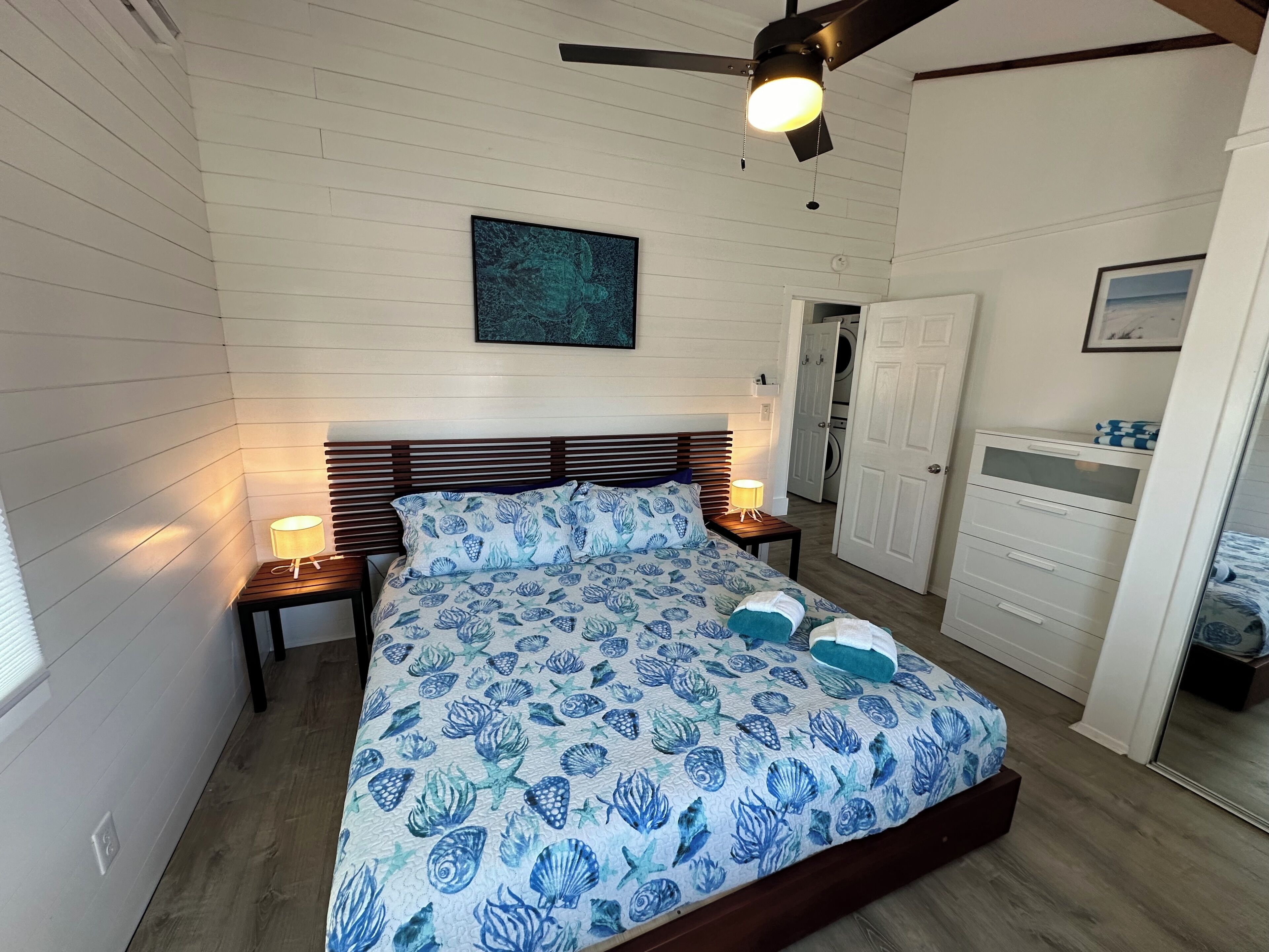 Een slaapkamer met een bed, houten hoofdbord, twee bedlampen, een plafondventilator, een aan de muur hangend kunstwerk, een kledingkast en een deur naar een andere kamer.