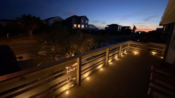 A wooden deck with railings and string lights, overlooking a residential area with houses and a sunset in the background. - Private Heated Pool and Hot Tub, across the street from the ocean! (Oak Island)