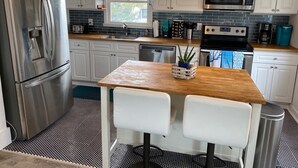 A modern kitchen with white cabinets, stainless steel appliances, and a wooden dining table with two chairs. - Private Heated Pool and Hot Tub, across the street from the ocean! (Oak Island)
