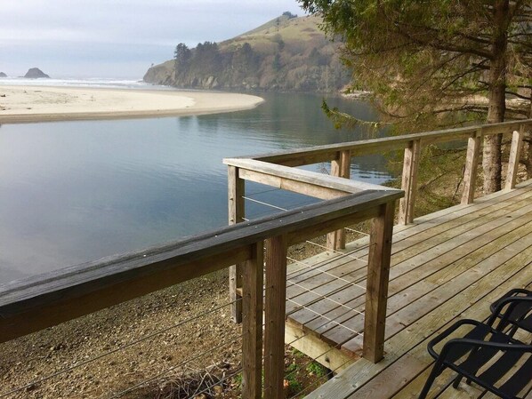 Een houten terras met uitzicht op een rustige baai, een zandstrand en heuvels in de verte.