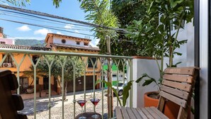 Een balkon met een houten bankje, twee kleine tafels met rode drankjes en een bloempot.