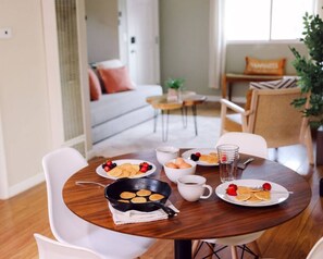 Un coin repas avec une table ronde en bois, des chaises blanches, des assiettes de crêpes garnies de fruits rouges, une tasse de café et une poêle.