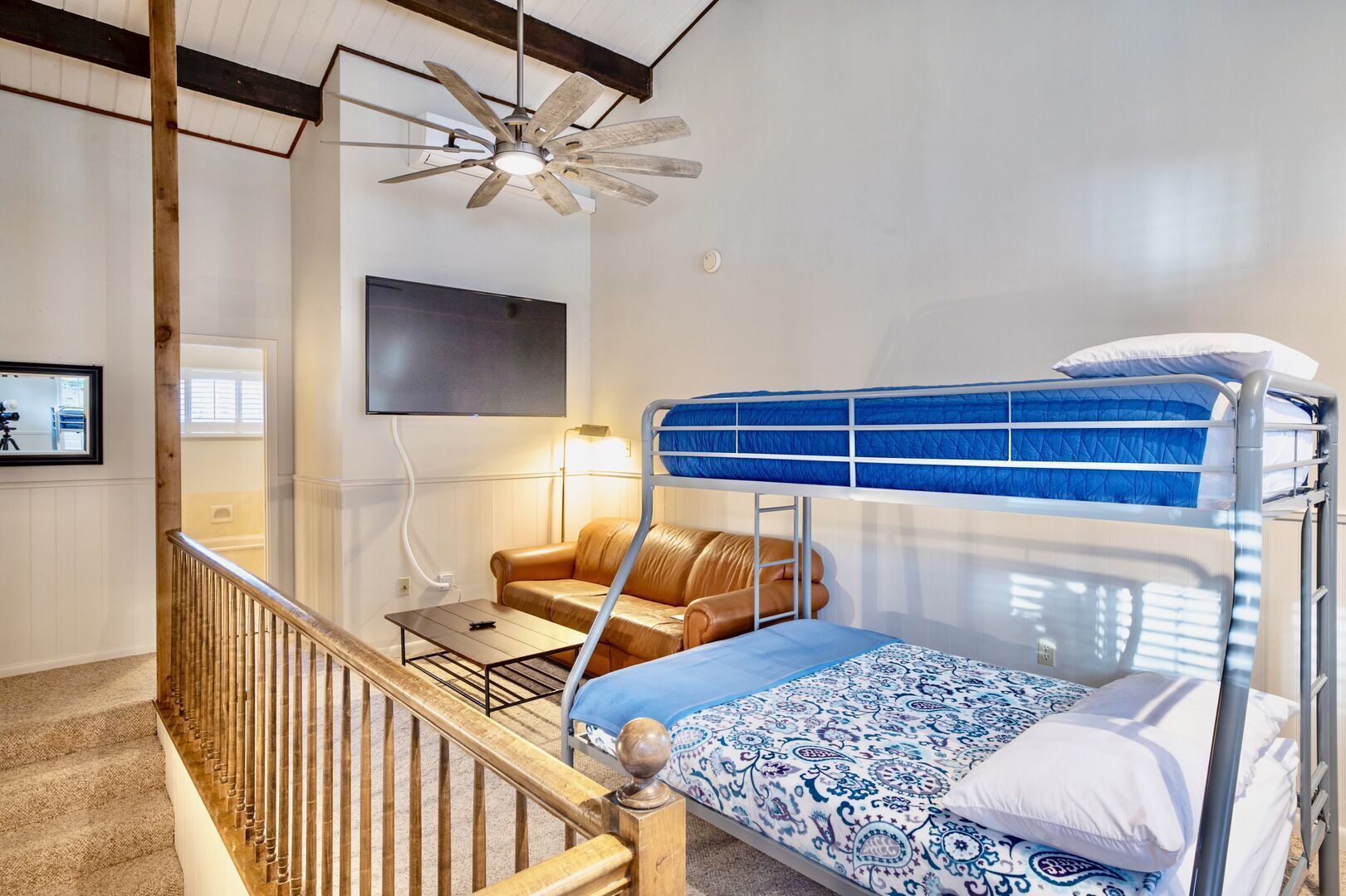 A bunk bed with a patterned bedspread, a wooden ladder, a leather sofa, a small table, and a ceiling fan.