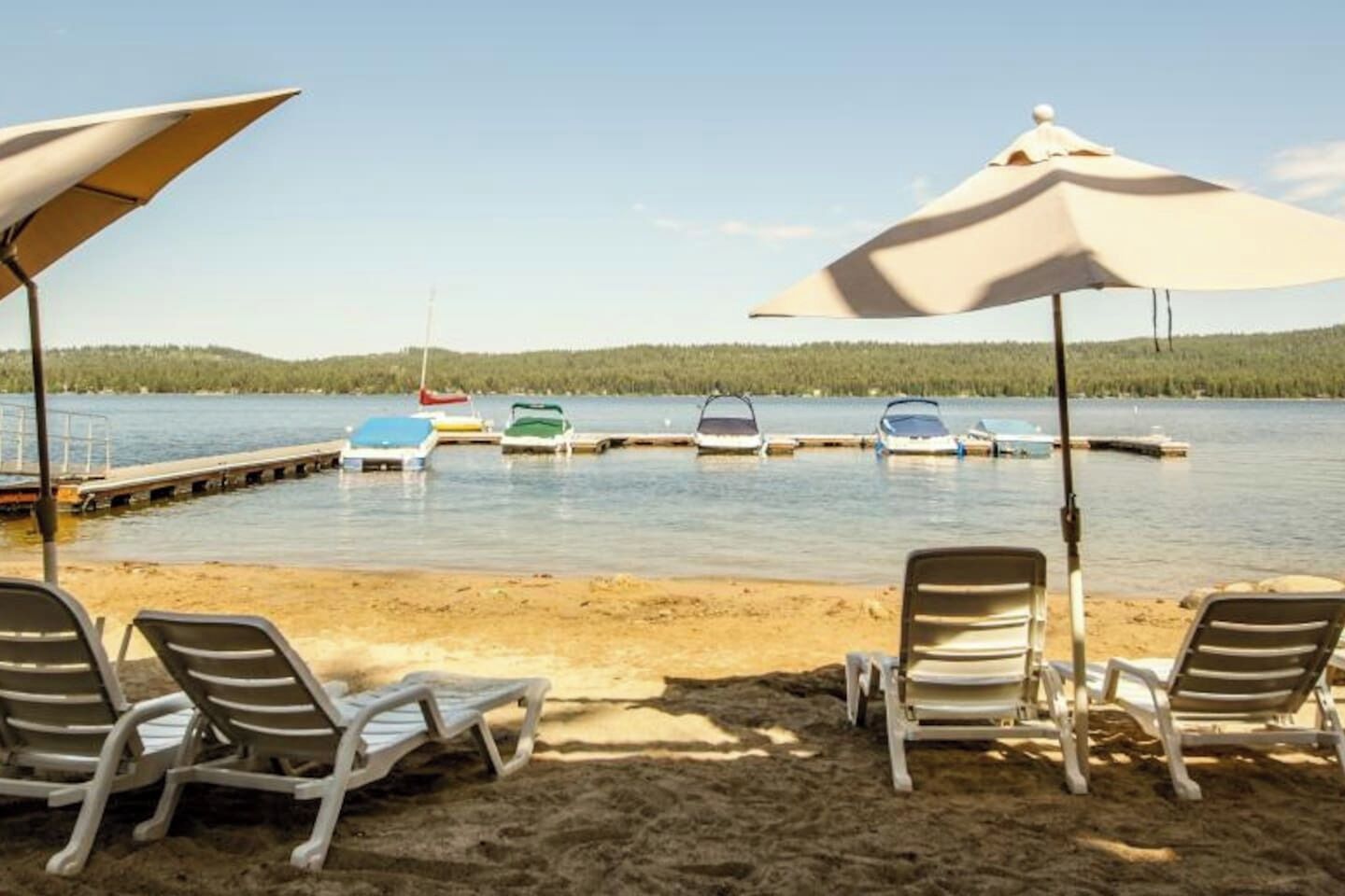 Uma praia à beira do lago com espreguiçadeiras e guarda-sóis, barcos atracados ao fundo, e uma colina arborizada distante.