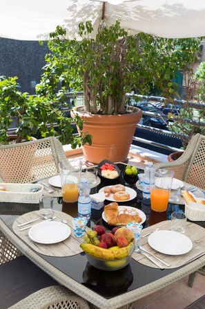 A breakfast table set with plates, cutlery, a fruit bowl, and beverages, surrounded by wicker chairs and a large potted plant.