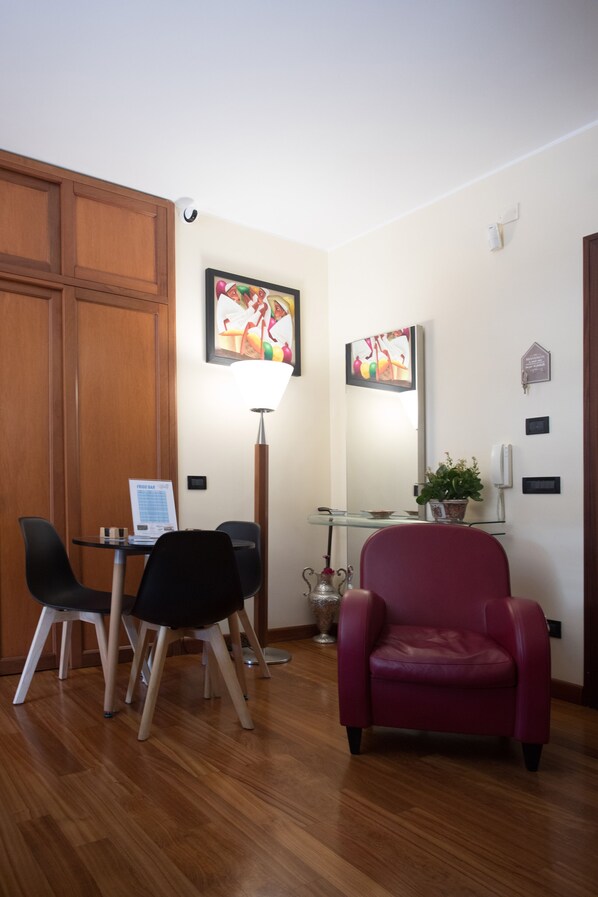 A room with a wooden door, a desk with a laptop, two black chairs, a red chair, a glass table, a floor lamp, a potted plant, and two framed artworks on the wall.