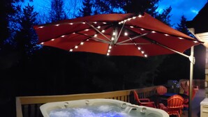 A hot tub with a red umbrella and string lights, surrounded by deck chairs and a wooden deck.