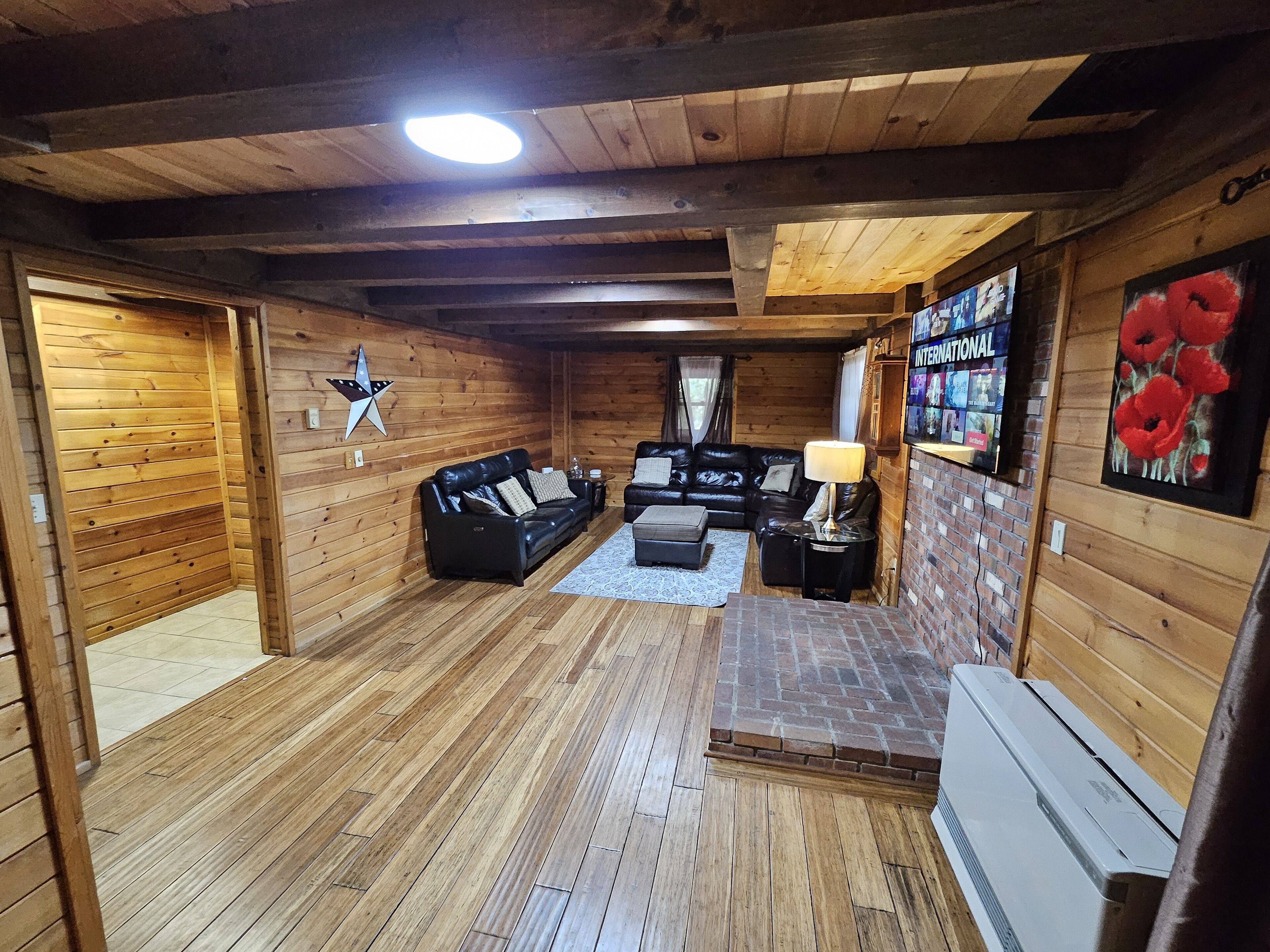 Ein Interieur einer Holzhütte mit Holzboden, einer schwarzen Ledersofa, einem Kamin und einem an der Wand befestigten Fernseher.