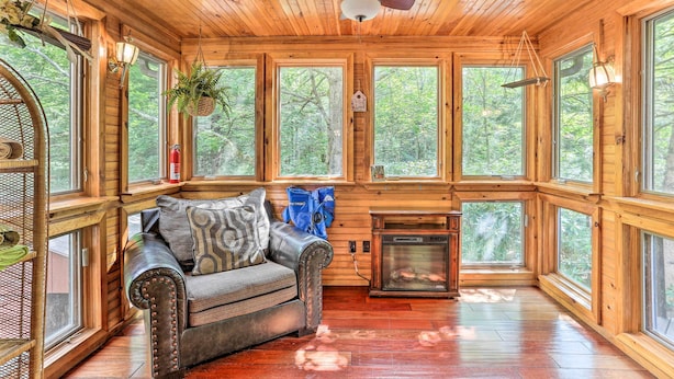 A cozy cabin interior with a wooden ceiling, large windows, a fireplace, and a cushioned seating area.