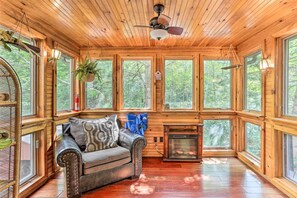 A cozy cabin interior with a wooden ceiling, large windows, a fireplace, and a cushioned seating area.