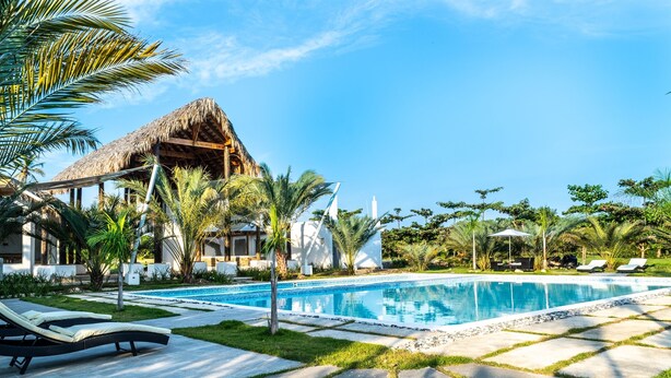 Un área de piscina con sillones de descanso, una estructura de techo de paja y vegetación tropical.