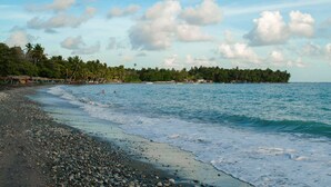Una playa con piedras, agua tranquila y una costa lejana con árboles y edificios.