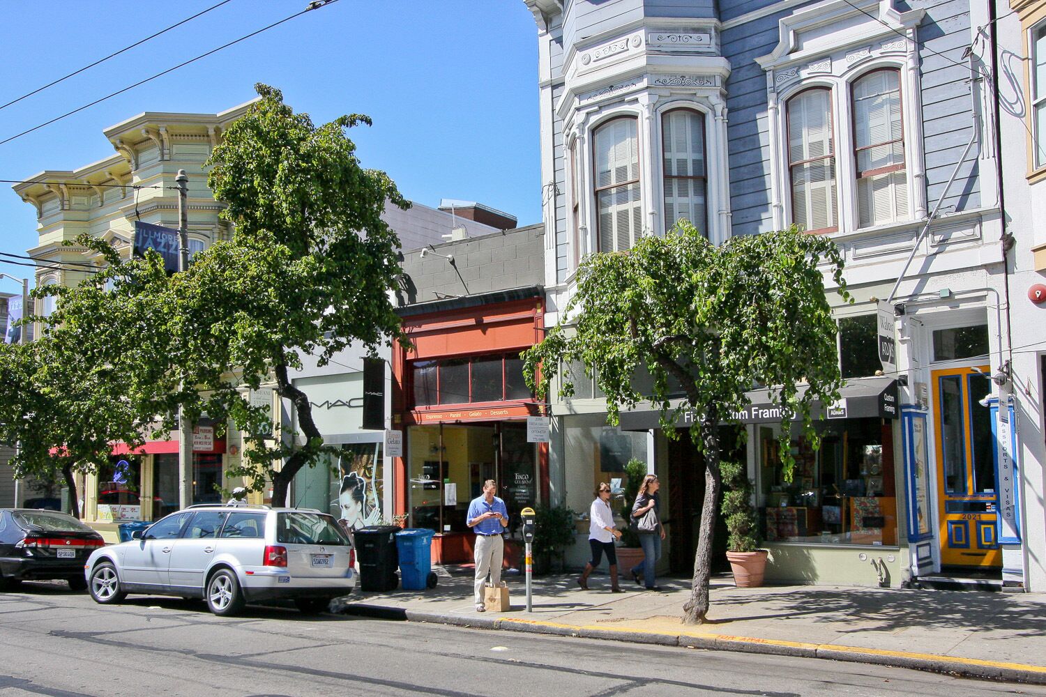 A street with parked cars, pedestrians, and buildings with commercial establishments.