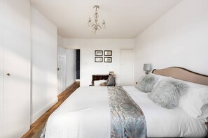 Une chambre à coucher avec un grand lit, un lustre, un bureau avec une chaise et des tableaux encadrés accrochés au mur.