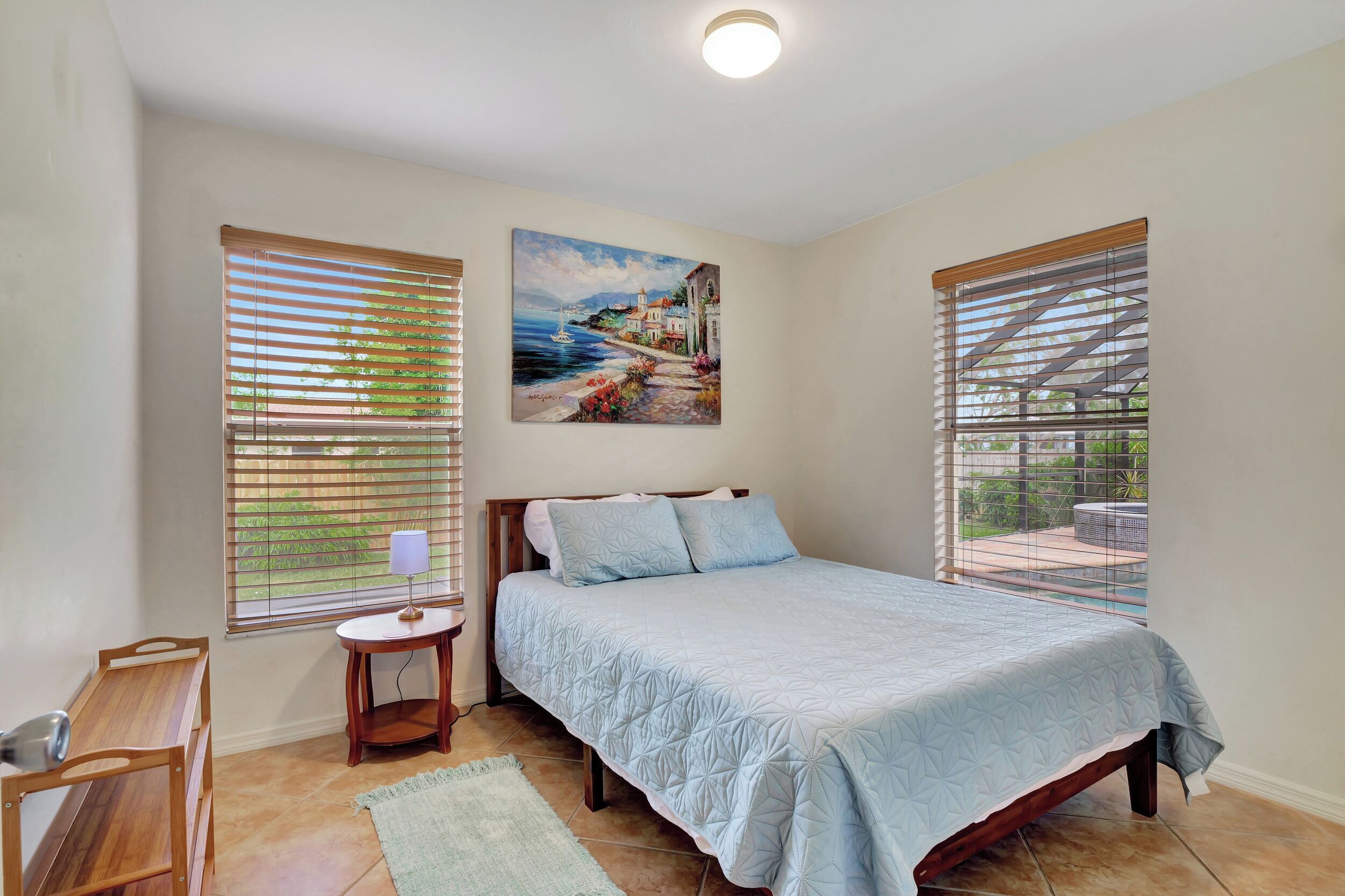 A bedroom with a bed, a nightstand, a lamp, a wooden chair, and a painting of a coastal scene.