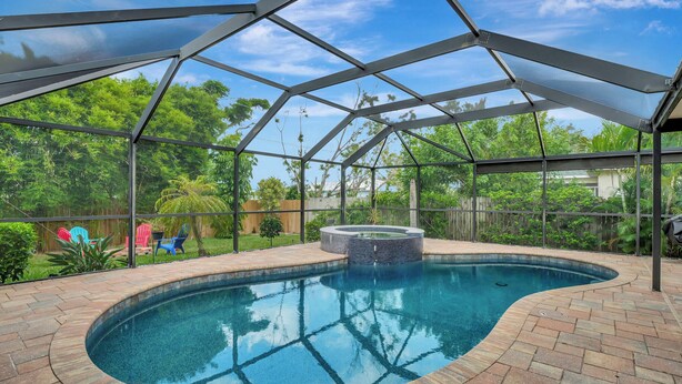A covered swimming pool with a hot tub, surrounded by a brick patio and a metal roof structure.