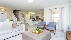 Un salon moderne avec un canapé, un pouf et deux fauteuils. Un escalier mène à la cuisine, que l’on aperçoit au fond.
