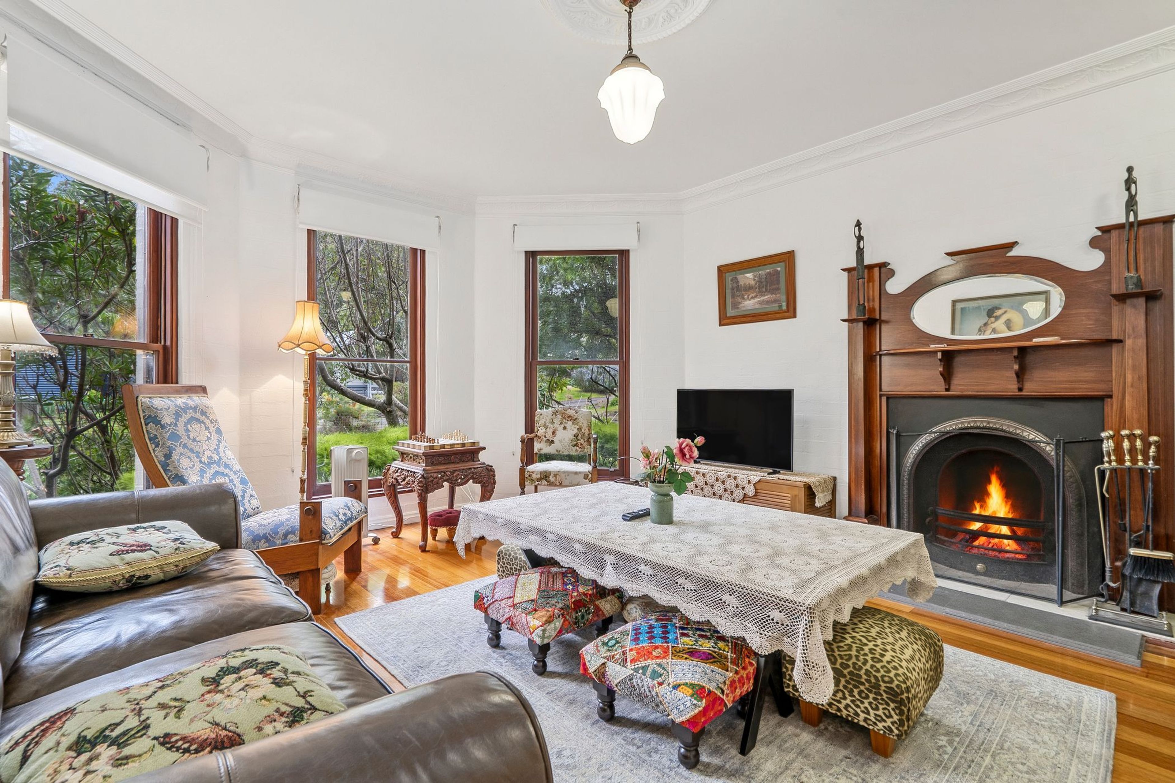 Ein traditionell eingerichtetes Wohnzimmer mit Kamin, einer Couch, einem Tisch mit weißer Spitzenleinentischdecke und Blick ins Freie durch große Fenster.