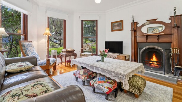 A traditional living room with a fireplace, a sofa, a table with a lace tablecloth, and a view of the outdoors through large windows.
