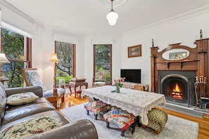 A traditional living room with a fireplace, a sofa, a table with a lace tablecloth, and a view of the outdoors through large windows.