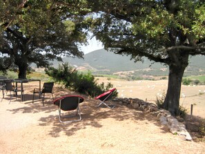 An outdoor seating area with chairs and a table under a tree, overlooking a valley with hay bales and mountains in the distance.