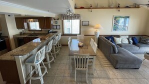 A spacious kitchen with a dining area, a gray sofa, and a large window. - Windhaven 301 - Oceanfront in Surfside Beach (Surfside Beach)
