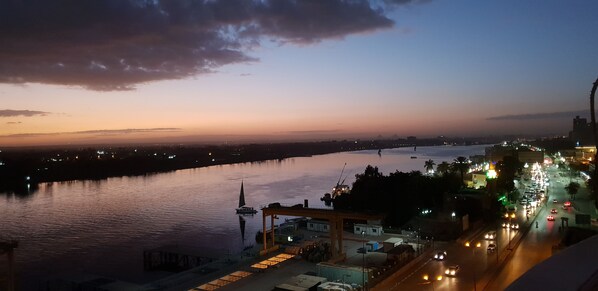 A river with a boat and a cityscape with buildings and a road. - Charming sunset,with Panoramic Nile view and pyramid view. (Cairo)