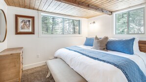 Un dormitorio con techo de madera, una cama grande con ropa de cama azul y blanca, dos ventanas y un aparador de madera.