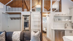 Un espacio habitable compacto con un dormitorio en un altillo, una kitchenette y una zona de estar con sofá y cojines.