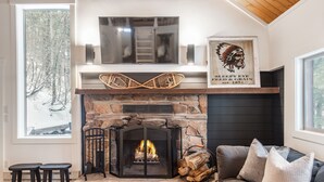 Un salón acogedor con chimenea de piedra, un sofá gris y un ventanal con vistas a un paisaje nevado.