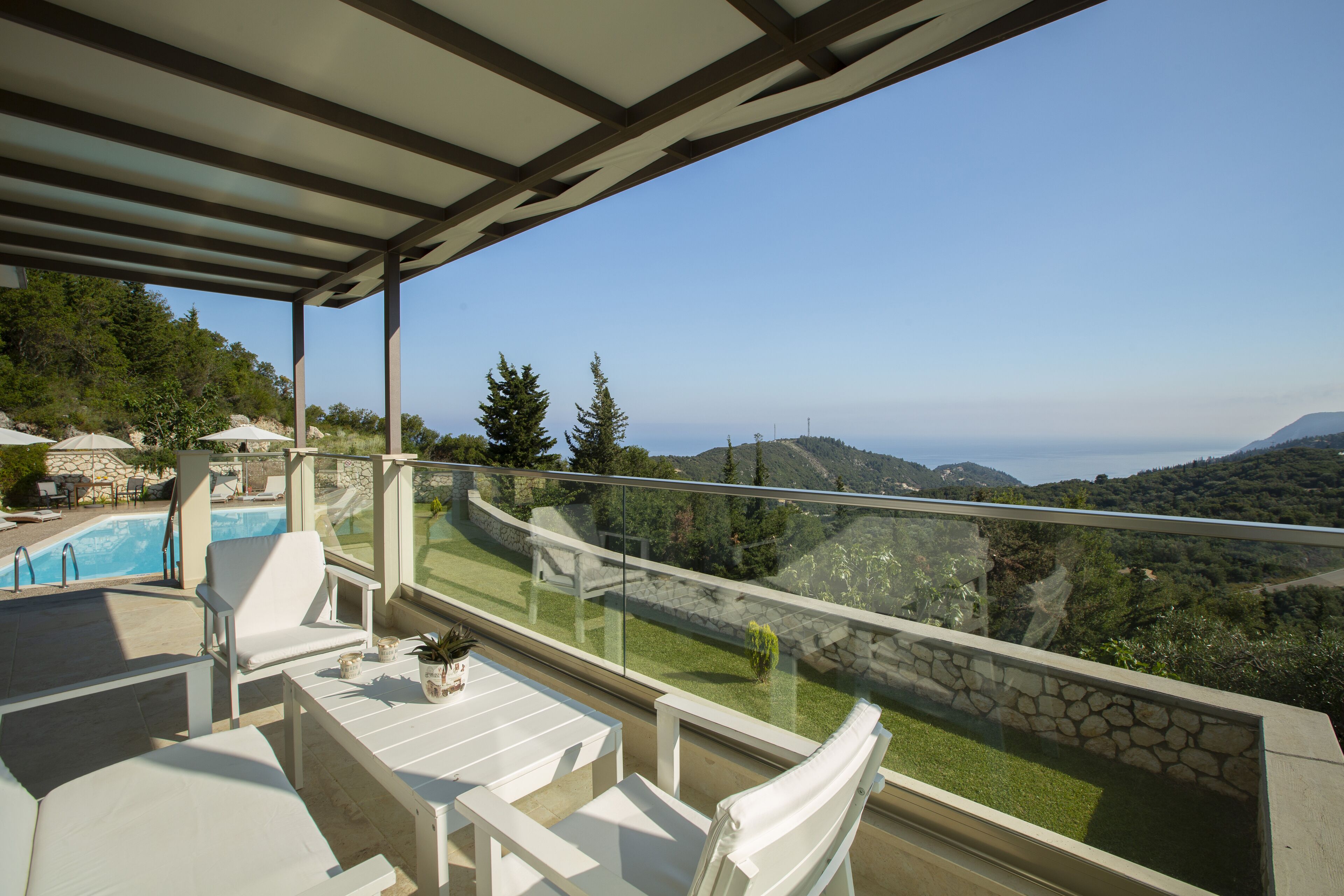 A balcony with white lounge chairs, a small pool, and a glass railing overlooking a mountainous landscape.