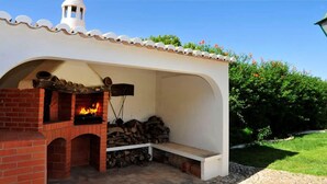 Forno in mattoni all'aperto con fuoco acceso, una panca e una pila di legna.